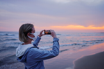 Traveling woman in medial mask taking photo of wonderful sunset on sea beach.