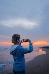 Traveling woman in medial mask taking photo of wonderful sunset on sea beach.