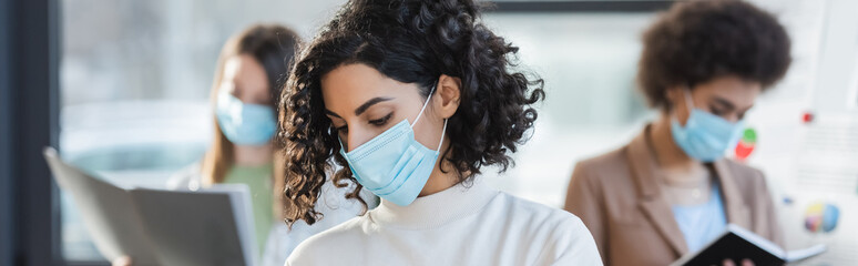 Muslim businesswoman in medical mask standing near blurred colleagues in office, banner.