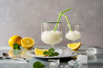 Lemon sorbet in two glasses on plate with lemons, ice cubes, mint leaves and drinking straws on grey background