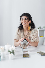 Cheerful gypsy medium touching orb near candles and tarot on table.