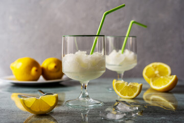 Lemon sorbet in two glasses with lemons, ice cubes and drinking straws on grey background