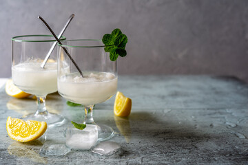Lemon sorbet in two glasses with lemons, ice cubes, tea spoons, mint leaves on grey background with copy space - obrazy, fototapety, plakaty