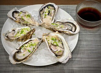 Six fresh Japanese large oysters with spring onions on top on a white plate with ice. A bowl of dipping sauce on the side.