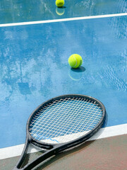 Black tennis racket and yellow tennis balls on wet tennis court with sunshine and reflections