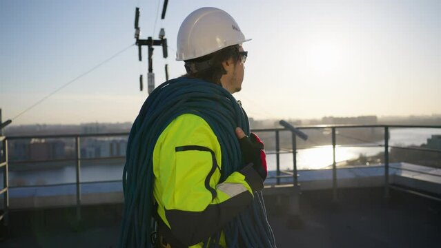 Climber in white helmet carries rope. Industrial climber with skein of rope on shoulder walks along rooftop of new building at sunset closeup