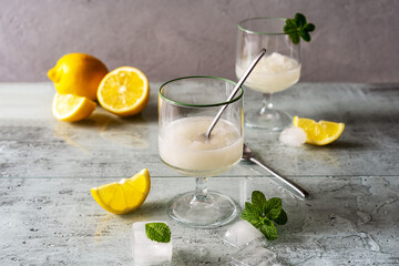 Lemon sorbet in two glasses with lemons, ice cubes, tea spoons, mint leaves on grey background