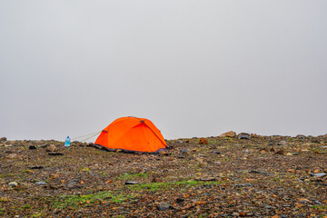Summer camping in top foggy mountain. Alpine landscape with orange tent at very high altitude in dramatic clouds. Awesome mountain scenery with tent above foggy in mountain.