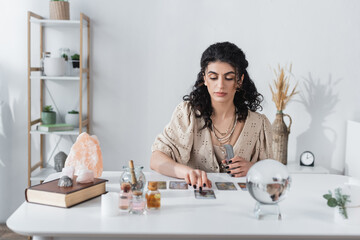 Gypsy fortune teller holding tarot cards near blurred orb on table.