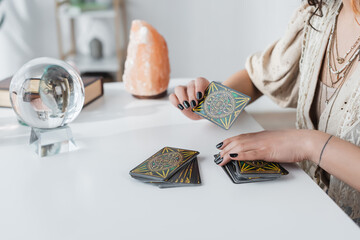 KYIV, UKRAINE - FEBRUARY 23, 2022: Cropped view of medium holding tarot cards near orb and blurred crystal on table. - obrazy, fototapety, plakaty