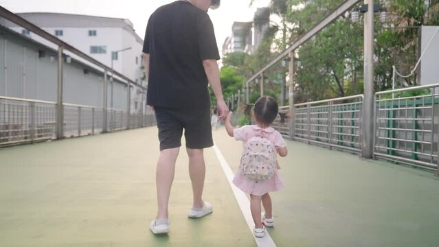 Young Asian father walking his daughter to school in the morning, family outdoor activity, daddy holding daughter hands, small cute child with school backpack walking along city pedestrian walkway