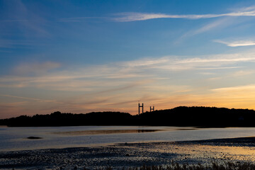 Sunset with reflections in water and silhouette of treeline. Bridge pylons in distance.