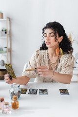 KYIV, UKRAINE - FEBRUARY 23, 2022: Young gypsy fortune teller holding tarot card and sage stick at...