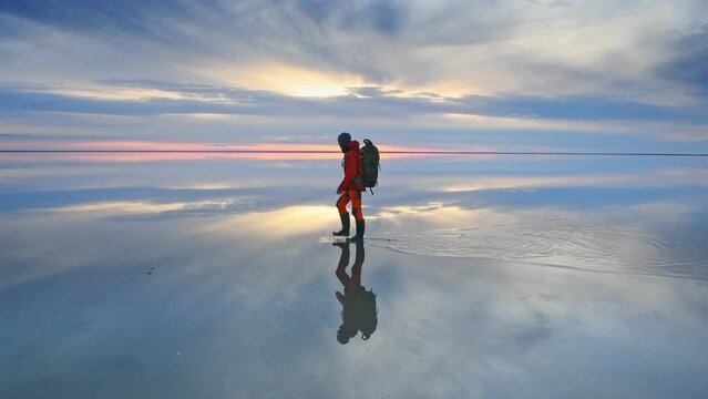Man traveler with backpack walking on the salt lake at sunset. Blue sky with clouds are reflected in the mirror water surface. Travel and adventure concept
