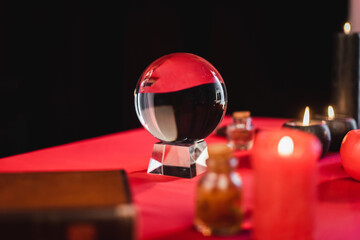 Glass orb near blurred candles and witchcraft supplies on table isolated on black.
