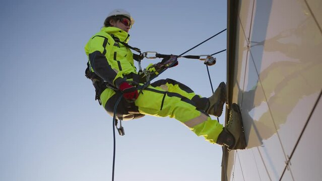 Industrial climber carefully climbs down from roof of skyscraper. Climber in safety equipment slowly moves down wall of highrise building