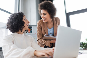 Smiling multiethnic businesswomen talking near laptop in office.