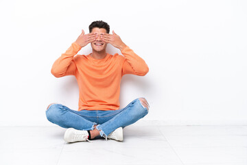 Young man sitting on the floor isolated on white background covering eyes by hands