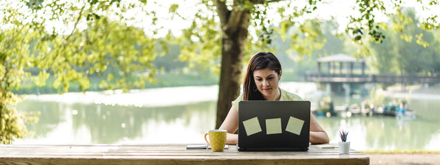 Horizontal banner with young woman using laptop computer in the public park - Millennial female freelancer working remotely and taking notes in a city park - Technology and remote work concept