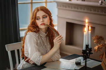 Portrait red-haired vintage woman writer holding bird feather in hands. Medieval girl sit at table writes book, letters on sheets paper with ink pen. Photo old retro style. White blouse, classic room - obrazy, fototapety, plakaty
