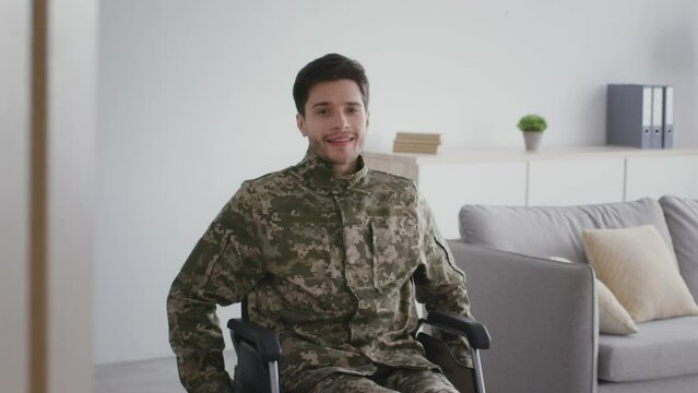 Young man soldier in camouflage with disability deeply breathing and smiling to camera, sitting in wheelchair at home