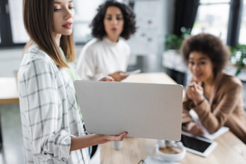 Fototapeta premium Laptop in hand of blurred businesswoman working with interracial colleagues in office.