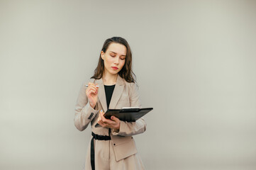 Beautiful woman in a jacket stands on a beige background with a serious face and writes in a paper tablet isolated.