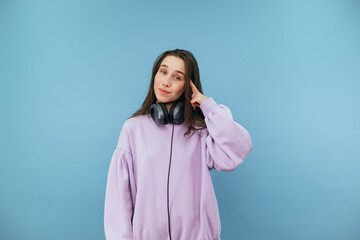 Beautiful woman in headphones and casual clothes points her finger at her temple and looks at the camera with a smile on her face on a blue background.