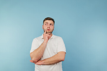 Pensive man in a white T-shirt stands on a blue background and looks away and thinks.