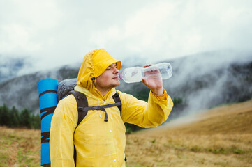 A male tourist in a raincoat and with a backpack on his back stands in the mountains and drinks water from a bottle against the background of a foggy mountain landscape.