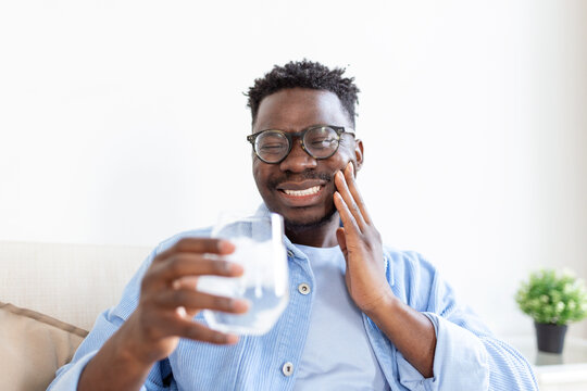 Young man with sensitive teeth and hand holding glass of cold water with ice. Healthcare concept. man drinking cold drink, glass full of ice cubes and feels toothache, pain