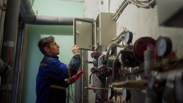 Electrician inspecting control panel with multimeter in a district heating substation, goes away and turns off light. Master in uniform checking fuse box with tester probes in technical room.