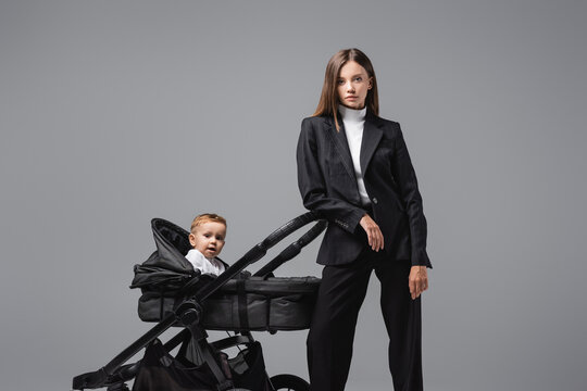 businesswoman in suit looking at camera near son in black pram isolated on grey.