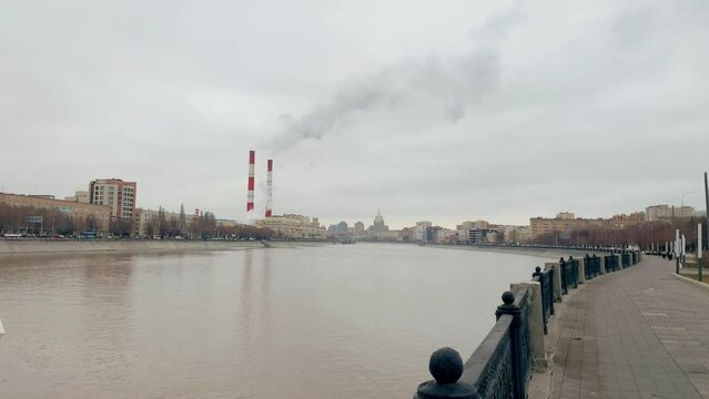 The embankment of the Moskva River, a walking area is visible on the right, a river, houses on the left, two white and red pipes are visible in front, from which smoke is coming. High quality 4k