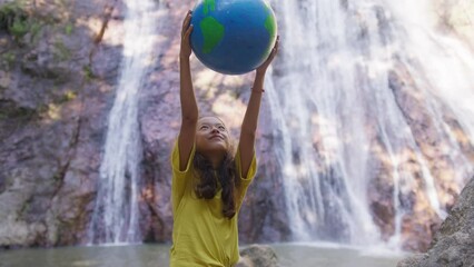 The girl stands in front of the waterfall, in nature, raises the globe above her head and examines it. World environment day concept, pollution, global warming, earth day.