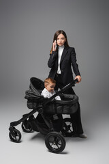 young woman calling on mobile phone near son in baby carriage on grey.