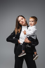 woman in black suit holding amazed son while standing with hand on hip isolated on grey.