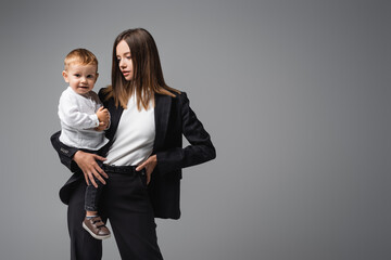 businesswoman standing with hand on hip while holding smiling son isolated on grey.