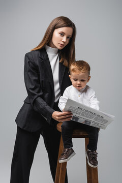 woman in black suit holding business newspaper near toddler son isolated on grey.