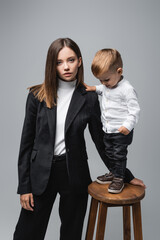 little boy standing on high stool near mother looking at camera isolated on grey.