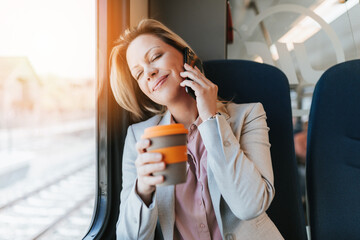 A middle-aged business woman is sitting in the cab of a high-speed train, looking through window and talking on phone.