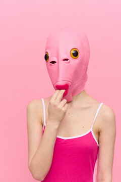 Funny woman in a sexy costume with a pink fish mask on her head put her fingers in her mouth in pink clothes on a pink background. The concept of modern art photography