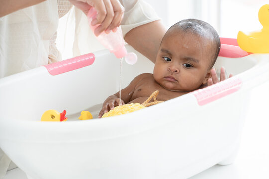 Happy adorable African newborn baby bathing in bathtub. Mother pouring shower gel on sponge to wash her little daughter in warm water. Newborn baby cleanliness care concept