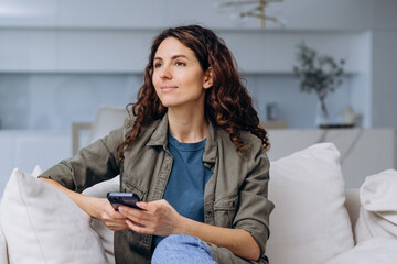 Happy young woman smiling holding smartphone in her hands while sitting on sofa at home and looking out the window, received a nice message or dreaming about vacation