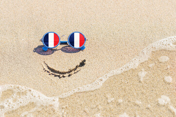 Fototapeta na wymiar A painted smile on the beach and sunglasses with the flag of France. The concept of a positive holiday in the resort of France.