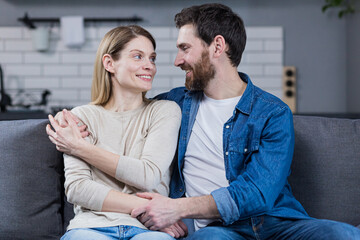 Portrait of a happy young family, couple. Man and woman sitting on sofa, hugging, waving, smiling, looking at camera, looking at each other