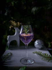 Vertical closeup of a glass of purple cocktail with rosemary and lemon on the table