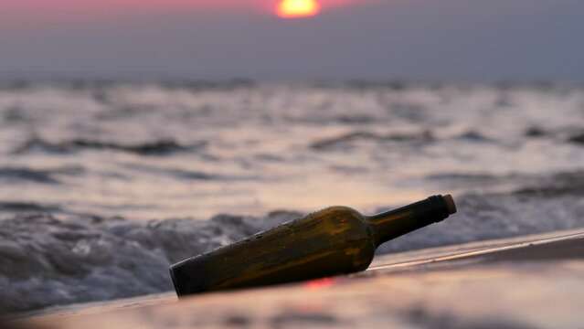 Bottle with message thrown ashore lies and then swayed from splash of oncoming wave. Close up shot of glass bottle traveled across sea to unknown beach, blurred sunset visible on background