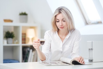 A young beautiful Caucasian blonde woman rests at home. A woman sits at a table, drinking coffee from a glass cup and reading a magazine.