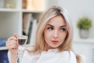 A young beautiful Caucasian blonde woman rests at home. A woman sits at a table, drinking coffee from a glass cup and reading a magazine.